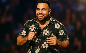 A man in a floral shirt smiles while holding a microphone on stage.