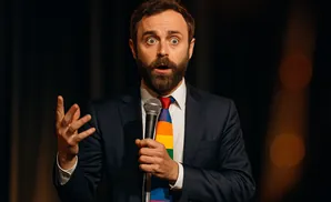 A man in a suit and rainbow tie holds a microphone, gesturing with his left hand.