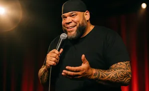 A man with a beard and tattoos on his arm, wearing a black t-shirt and baseball cap, smiles while holding a microphone.