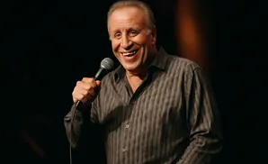 A man in a striped shirt laughs while holding a microphone.
