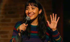 A woman with dark hair and bangs smiles as she holds a microphone and raises her hand.