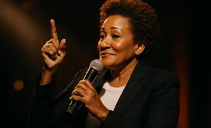 A Black woman in a black blazer speaks into a microphone, holding her index finger up in a 