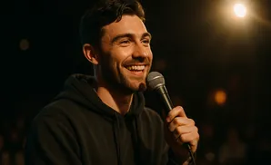 A man in a black hoodie smiles and holds a microphone, with a spotlight on him.