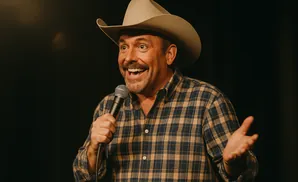 Comedian in a cowboy hat and plaid shirt, holding a microphone and gesturing with his other hand.