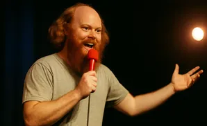 A man with red hair and beard sings into a microphone on a dark stage with a spotlight.
