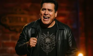 Comedian Carlos Mencia performs on stage, holding a microphone and wearing a black leather jacket.