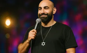 A bald, bearded man in a black t-shirt holding a microphone and wearing a Star of David necklace.