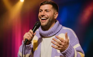 A man in a lavender sweater with a flower pattern holds a microphone and smiles warmly on a stage with colorful lights.