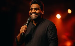 Comedian holding a microphone and laughing on a stage with orange lighting.