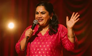 A woman in a pink polka-dot outfit smiles and waves while holding a microphone.