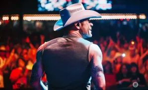 Man in a cowboy hat and tank top performs on stage with a crowd in the background.