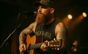 Man with a beard and tattoos plays an acoustic guitar on stage, lit by warm spotlights.