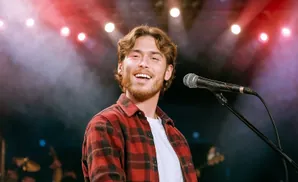 Young man with beard in red flannel shirt singing into a microphone on stage with red and white lights.