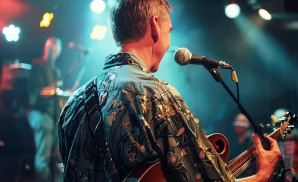 A musician in a patterned shirt plays guitar on stage with a microphone.