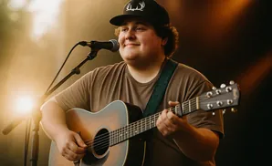 A young man wearing a baseball cap plays an acoustic guitar and sings into a microphone.