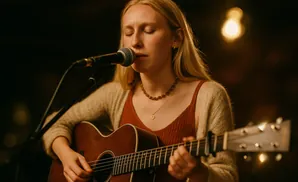 A woman with blonde hair plays an acoustic guitar and sings into a microphone.