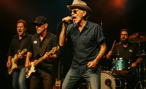 A band performing live with a singer in a hat, bassist, guitarist, and drummer on stage.