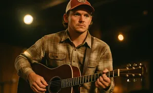 A man in a plaid shirt and baseball cap plays an acoustic guitar.