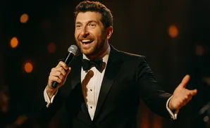 A man in a tuxedo holds a microphone and speaks into it with his mouth open and a hand outstretched.