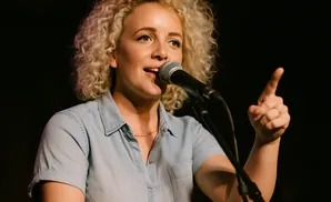 Woman with curly blonde hair, wearing a blue denim shirt, speaking into a microphone and pointing her finger.