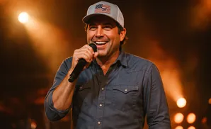 A man in a denim shirt and baseball cap sings into a microphone on a stage, lit by warm spotlights.