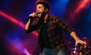 A male singer with a beard in a plaid shirt sings into a microphone with stage lights in the background.