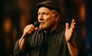 A man in a cap and dark shirt speaks into a microphone with his hand raised.