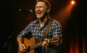 A man in a plaid shirt plays an acoustic guitar on stage, smiling into a microphone under warm stage lights.