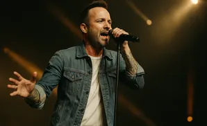 A man in a denim jacket sings into a microphone with his hand outstretched.