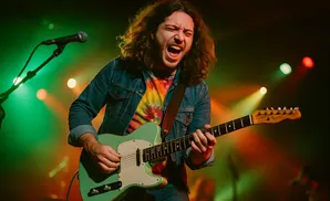 A male guitarist with long, curly hair wearing a denim jacket and tie-dye shirt plays a teal electric guitar on stage.