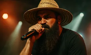 Man with a beard and straw hat singing into a microphone on a dimly lit stage.