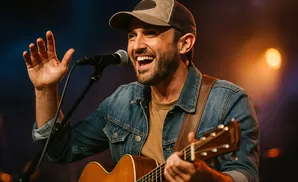 A man in a denim jacket and baseball cap sings into a microphone while playing an acoustic guitar on stage with stage lighting.