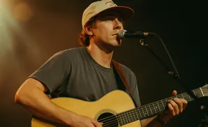 A man wearing a baseball cap plays an acoustic guitar and sings into a microphone.