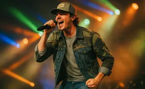 A male singer performs on stage wearing a camouflage jacket and a baseball cap, illuminated by colorful spotlights.