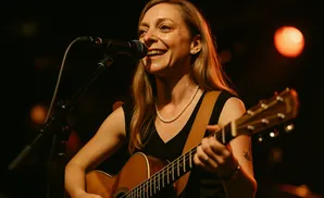 A woman sings into a microphone while playing an acoustic guitar on stage.