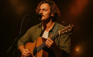 A man with curly hair plays an acoustic guitar and sings into a microphone on a dimly lit stage.