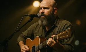 Bald man with a beard playing an acoustic guitar and singing into a microphone.