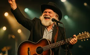 A joyful, white-bearded man in a cowboy hat sings and plays guitar under stage lights.