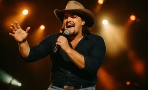 A male country singer in a cowboy hat performs on stage, holding a microphone and smiling.