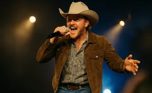 A male country singer in a cowboy hat and brown jacket performs on stage, holding a microphone.