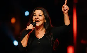 A woman in a black shirt sings into a microphone with her arm raised and finger pointing up.