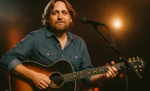 Musician playing an acoustic guitar and singing into a microphone, bathed in warm stage lighting.