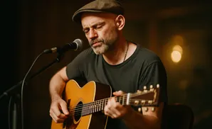 A man wearing a cap plays an acoustic guitar and sings into a microphone in a dimly lit room.