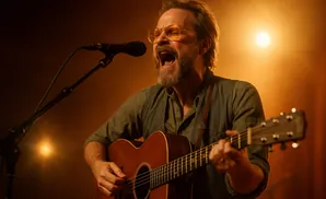 Man with beard wearing glasses and a grey shirt playing an acoustic guitar and singing into a microphone.