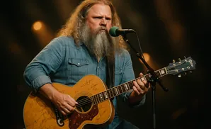 A man with long hair and a beard plays an acoustic guitar while singing into a microphone.