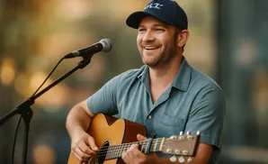 A smiling man in a blue shirt and baseball cap plays an acoustic guitar in front of a microphone.