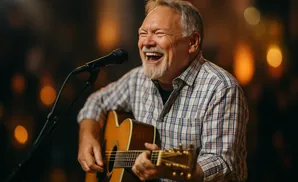 A smiling man with a grey beard plays an acoustic guitar and sings into a microphone.