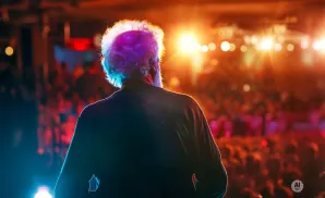 Man with white, curly hair on stage facing a blurred audience under bright lights.
