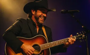 A man in a cowboy hat plays an acoustic guitar and smiles into a microphone on stage.