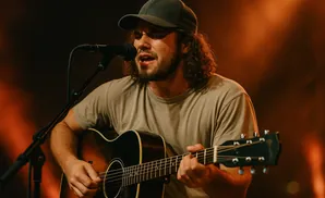 A man with curly hair and a baseball cap plays an acoustic guitar and sings into a microphone.